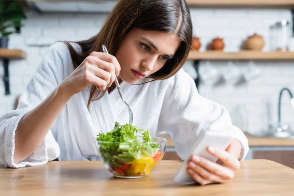 Upset young adult woman in bathrobe eating vegetables salad and using cellphone in modern kitchen — Stock Photo