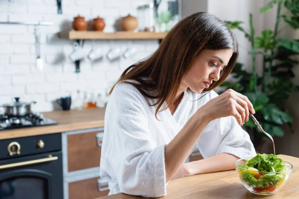 Traurige junge erwachsene Frau im Bademantel isst Gemüsesalat in moderner Küche — Stockfoto