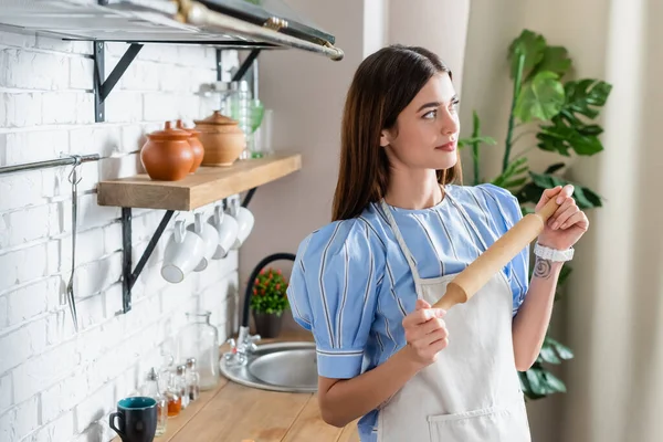 Вдумчивая взрослая женщина в фартуке, стоящая со скалкой из теста на современной кухне — стоковое фото