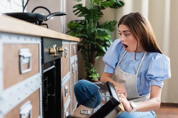 Amazed young adult woman in apron looking into oven with open mouth in modern kitchen — Stock Photo