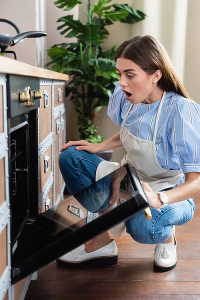 Erstaunte junge erwachsene Frau in Schürze blickt in moderne Küche mit offenem Mund in den Ofen — Stockfoto