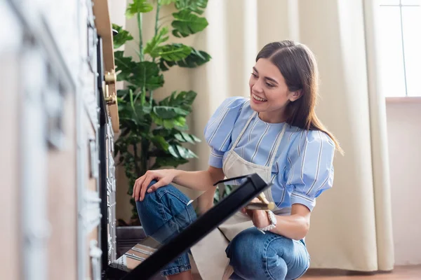 Sonriente joven mujer adulta en delantal mirando en el horno en la cocina moderna - foto de stock