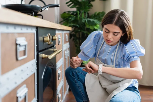 Молодая взрослая женщина в фартуке сидит рядом с духовкой и смотрит на наручные часы на современной кухне — стоковое фото