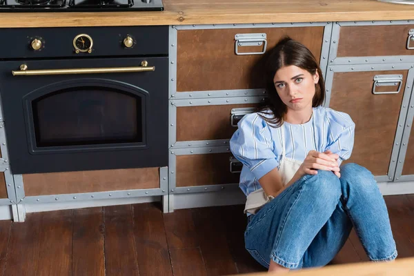 Cansada mujer joven adulta sentada en el suelo cerca del horno con en la cocina moderna - foto de stock