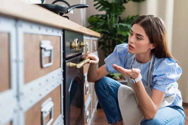 Young adult woman in apron looking at timer on oven and gesturing with hand in modern kitchen — Stock Photo