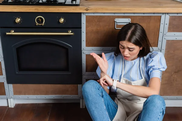 Смущенная молодая женщина в фартуке сидит рядом с духовкой и смотрит на наручные часы на современной кухне — стоковое фото