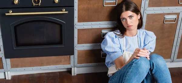 Upset young adult woman sitting on floor near oven with in modern kitchen, banner — Stock Photo