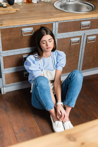 Fatigué jeune femme adulte assis sur le sol avec les yeux fermés dans la cuisine moderne — Photo de stock