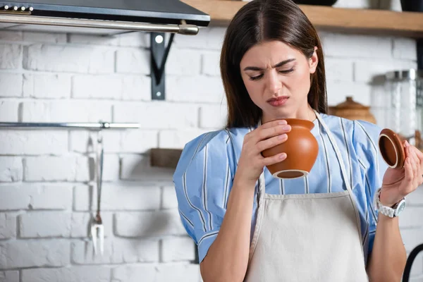 Junge erwachsene Frau in Schürze blickt angewidert auf Tontopf in Küche — Stockfoto