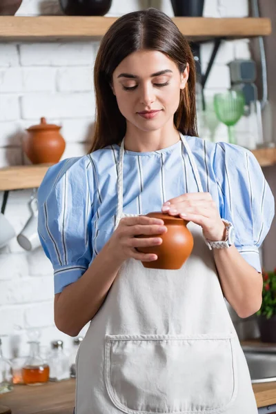 Alegre joven mujer adulta en delantal sosteniendo la olla de arcilla en las manos en la cocina - foto de stock