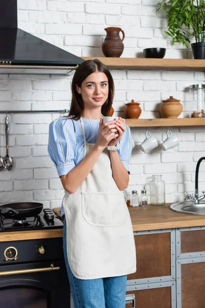 Улыбающаяся молодая женщина держит чашку кофе в руках на современной кухне — стоковое фото