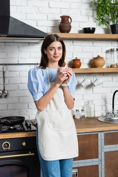 Sonriente mujer adulta joven sosteniendo la taza de café en las manos en la cocina moderna - foto de stock