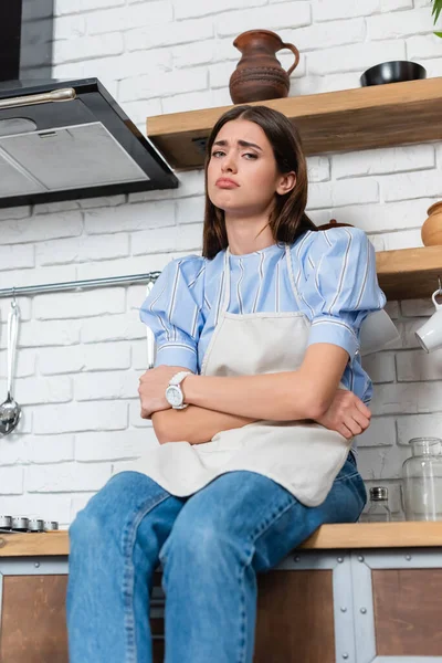 Triste joven mujer adulta en delantal sentado con los brazos cruzados en la cocina — Stock Photo