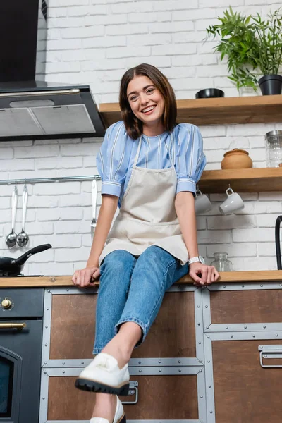 Happy young adult woman in apron sitting in modern kitchen — Stock Photo