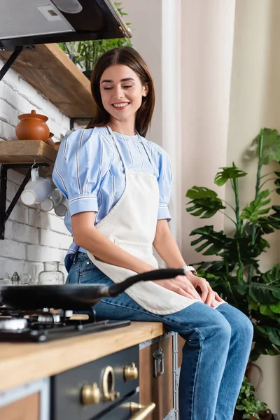 Smiling young adult woman in apron sitting near frying pan in modern kitchen — Stock Photo