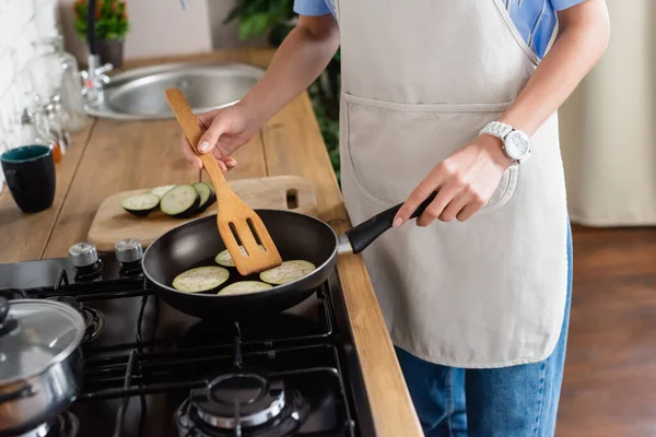 Cropped view of young adult woman frying slices of eggplant in pan and using spatula in kitchen — Stock Photo