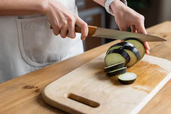 Vista de cerca de las manos femeninas de corte de berenjena en la tabla de cortar en la cocina - foto de stock