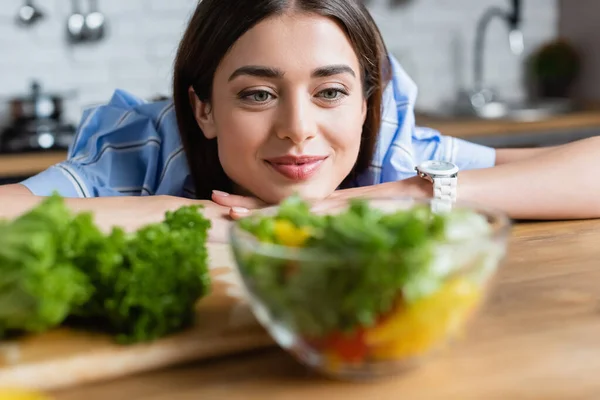 Sorrindo jovem mulher adulta olhando para preparado salada de legumes na cozinha — Fotografia de Stock