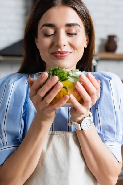 Souriant jeune femme adulte profitant de l'odeur de salade de légumes frais avec les yeux fermés dans la cuisine — Photo de stock