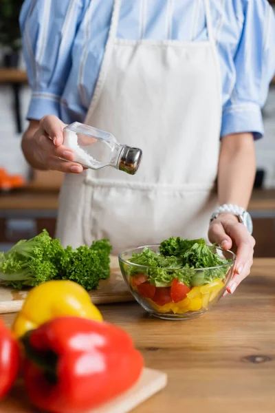 Partial view of young adult woman in apron seasoning fresh vegetables salad with salt mill in modern kitchen — Stock Photo