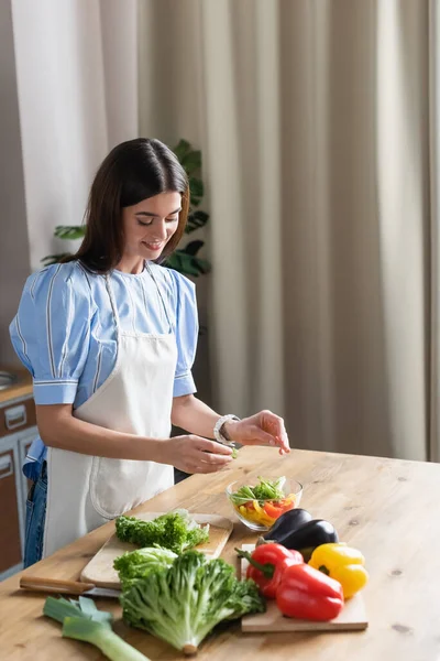 Sonriente mujer joven adulta en delantal cocinar verduras frescas ensalada en la cocina - foto de stock