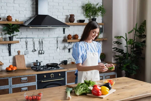 Happy young adult woman in apron preparing vegetables salad in modern kitchen — Stock Photo