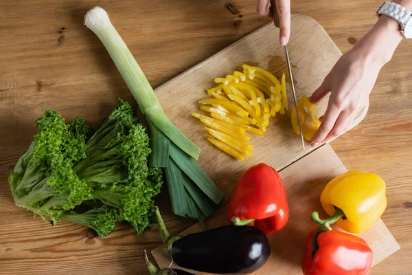 Ansicht von oben: Frauenhände hacken gelben Paprika auf Schneidebrett mit anderem Gemüse auf dem Tisch in der Küche — Stockfoto
