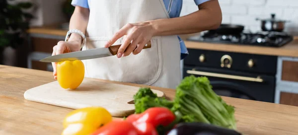 Partial view of young adult woman cutting yellow pepper on cutting board in kitchen, banner — Stock Photo