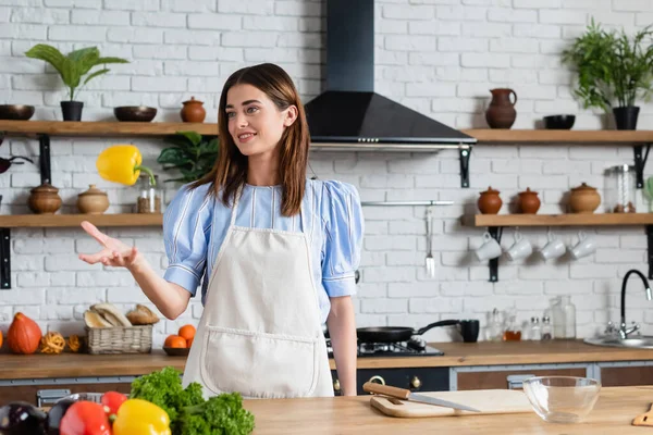 Attractive young adult woman throwing yellow pepper into air in kitchen — Stock Photo