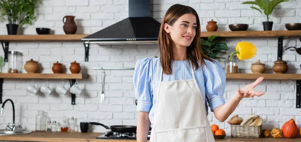 Attractive young adult woman throwing yellow pepper into air in kitchen, banner — Stock Photo