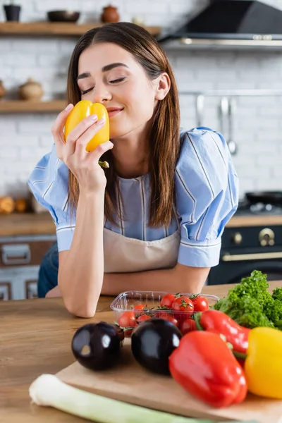 Smiling young adult woman smelling fresh yellow pepper in kitchen — Stock Photo