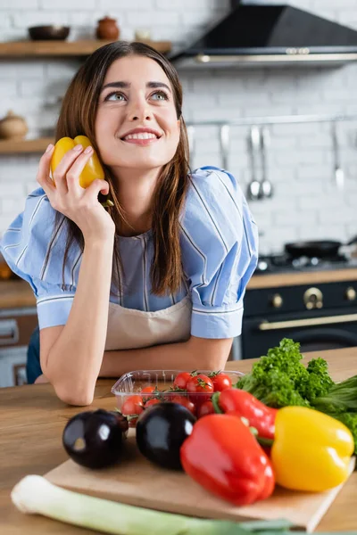 Mujer adulta joven soñadora sosteniendo pimienta amarilla fresca en la mano en la cocina - foto de stock