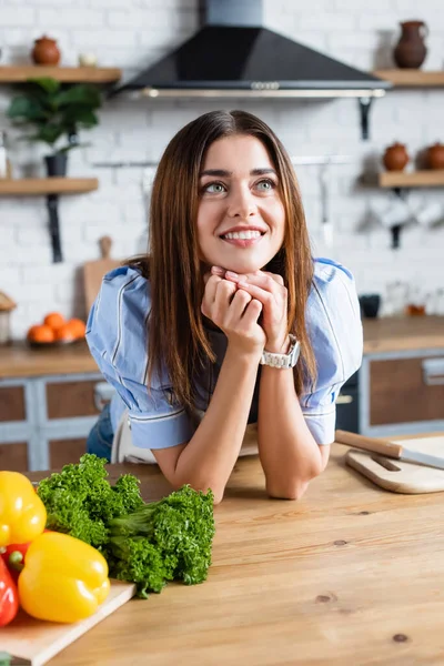 Joven mujer adulta de pie cerca de la mesa con verduras y cogidas de la mano cerca de la cara en la cocina - foto de stock