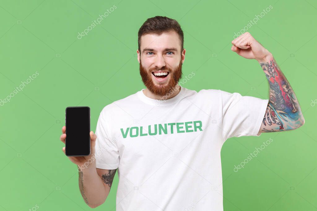 Strong man in volunteer t-shirt isolated on pastel green background. Voluntary free work assistance help charity grace teamwork concept. Hold mobile phone with empty screen, showing biceps, muscles
