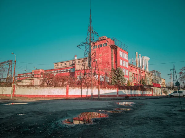 手前にはプドルと放棄された道路のあるターコイズブルーの空に対する赤い工業ビル — ストック写真