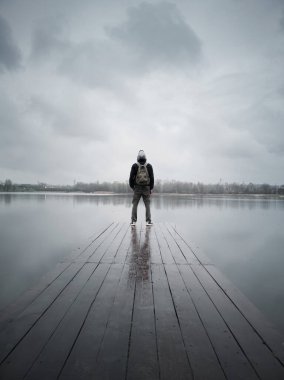Modern bir adam sırtı kameraya dönük durur, çerçevenin ortasında, bulutlu bir günde gölün üzerindeki küçük ahşap bir köprü iskelesinde..