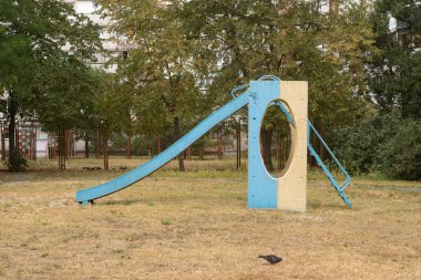 Eski mavi ve sarı beton parktaki kaydırak apartmanların yakınındaki kuru çimlerin üzerinde. Troyeshyna, Kyiv, Ukrayna, 7 Eylül 2025.