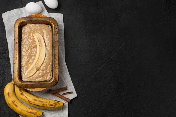 Raw banana bread dough in rectangular baking dish with ingredients on black background