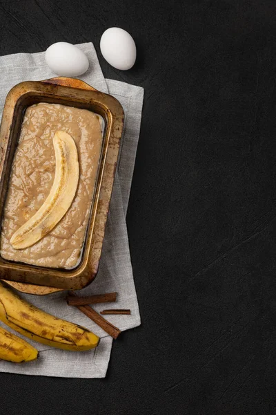 Raw banana bread dough in rectangular baking dish with ingredients on black background