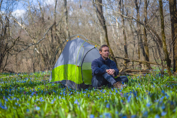 hiker sit near touristic tent on a forest glade with flowers, travel hiking background