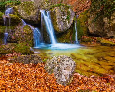 Nehirdeki şelale dağ kanyonu boyunca akar. Kırmızı kuru yapraklarla kaplıdır. Sonbahar nehir manzarası.
