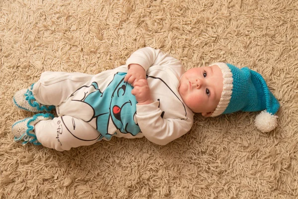 Baby in blue hat, lying on the beige carpet — Stock Photo, Image