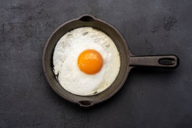 Bir yumurta, siyah taş arka planda dökme demir bir tavada pişiyor.