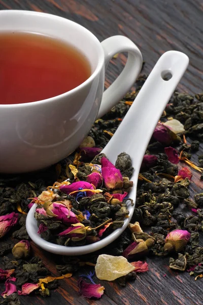a cup of green tea and dry tea leaves with flower petals in a spoon on a wooden table