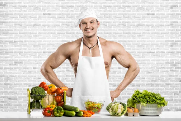 白いトックのブランチで男のボディビルダーと調理保護エプロン 調合野菜や笑顔 白いレンガの壁の背景に — ストック写真