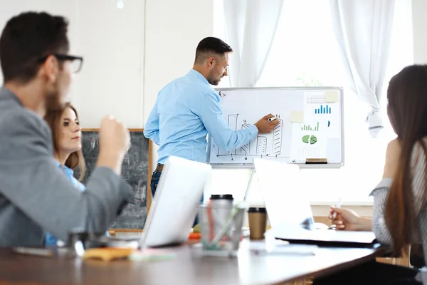 Schöner junger Mann steht neben Whiteboard und zeigt auf das Diagramm, während seine Kollegen zuhören und am Tisch sitzen — Stockfoto