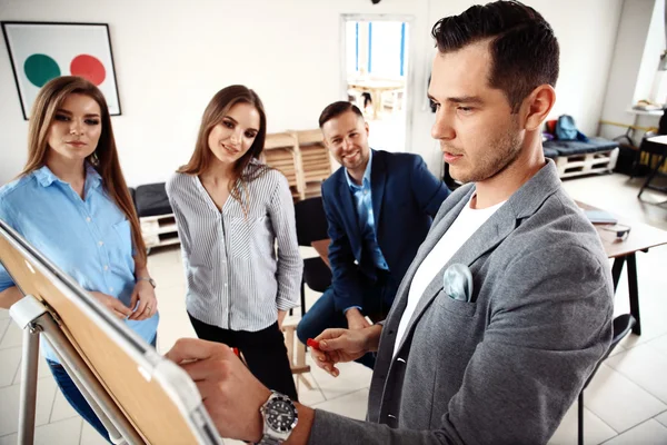 Knappe jonge man in glazen staan in de buurt van Whiteboard en wijzend op de kaart terwijl zijn collega's luisteren en zittend aan tafel — Stockfoto