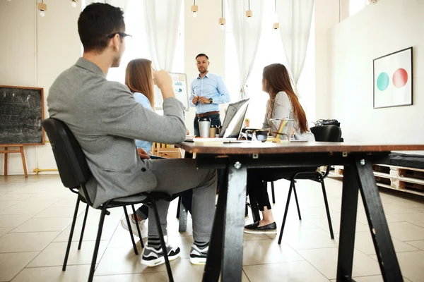 Handsome young man standing near whiteboard and pointing on the chart while his coworkers listening and sitting at the table — Stock Photo, Image