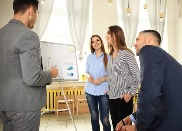 Handsome young man standing near whiteboard and pointing on the chart while his coworkers listening and sitting at the table — Stock Photo, Image