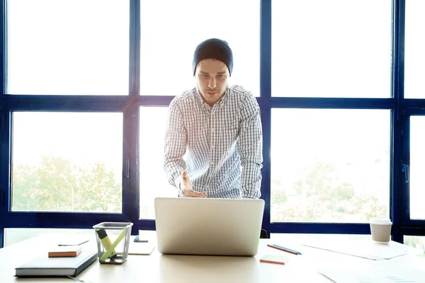 Nachdenklicher junger gutaussehender Mann arbeitet am Laptop, während er an seinem Arbeitsplatz sitzt — Stockfoto